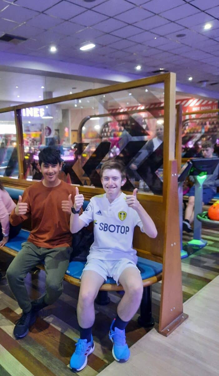 Two boys sit on a bench at a bowling alley looking at the camera smiling with their thumbs up.