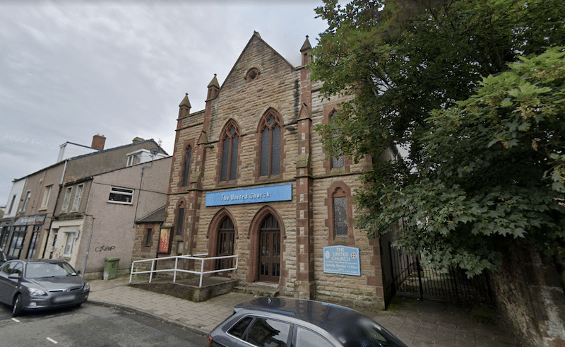 Stone church building, United Church Workington, with trees on the right and shops to the left.