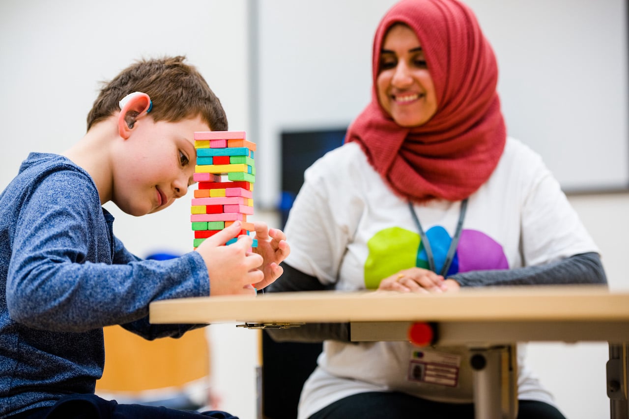 A boy with a hearing aid is sitting at the table playing a game, carefully removing bricks from a stack. A member of staff/volunteer is smiling as she watches.