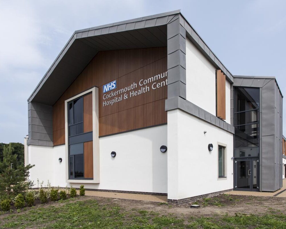 A modern building with white walls, wood panelling, big glass windows and grey around the windows, roof and door. The words 'NHS Cockermouth Community Hospital & Health Centre' are on the end of the building.