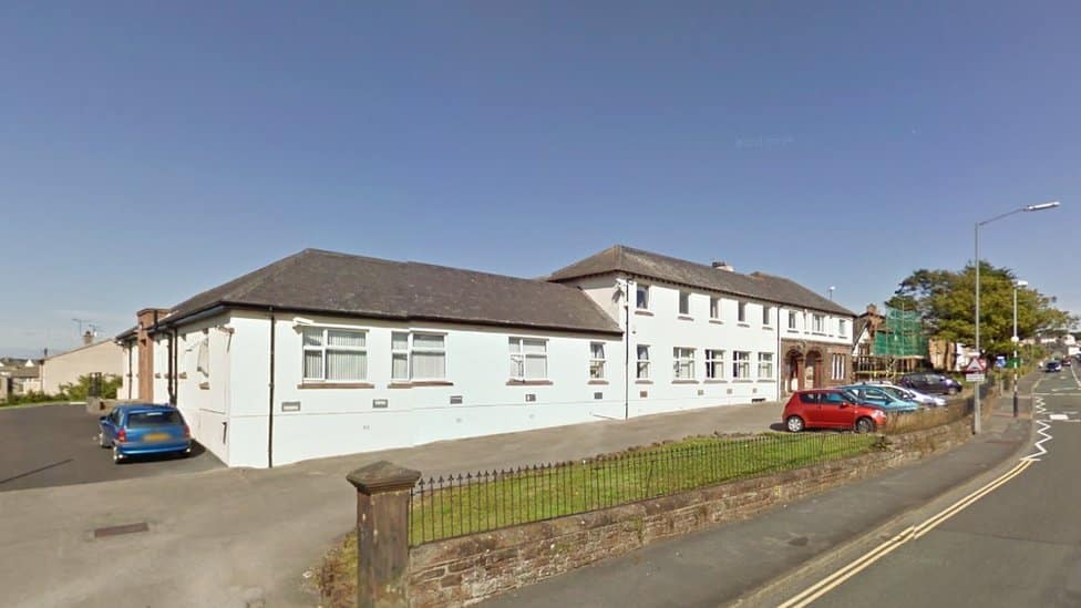 A white one storey building, leading into a two storey building with a car park wrapped around it, Maryport Cottage Hospital.