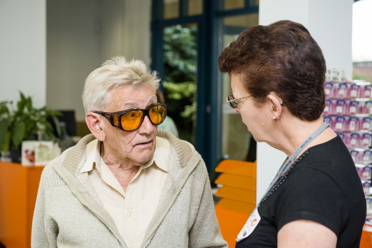 An elderly man is wearing yellow tinted glasses and is in conversation with a woman.