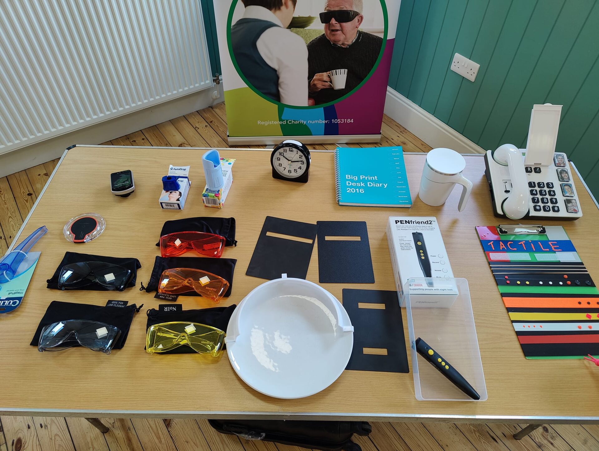 A table covered in VI equipment such as noir glasses, eye drop dispensers, big print diaries, alarm clock and tactile tester strips. 