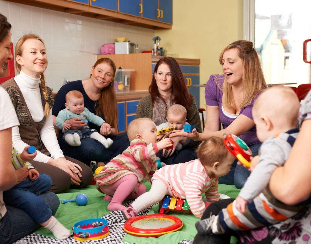 Group of 6 women sat in a circle with their babies either on their laps on crawling around on the floor between them.