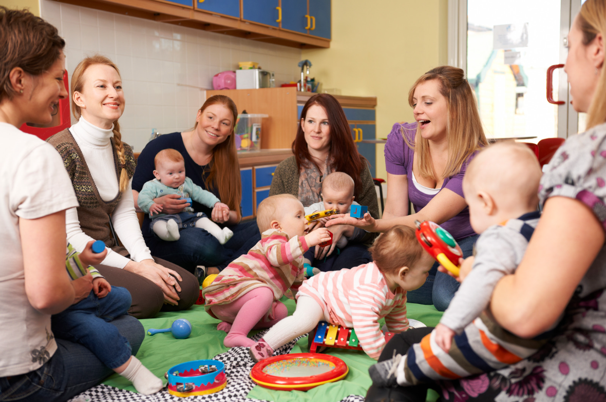 Group of 6 women sat in a circle with their babies either on their laps on crawling around on the floor between them.