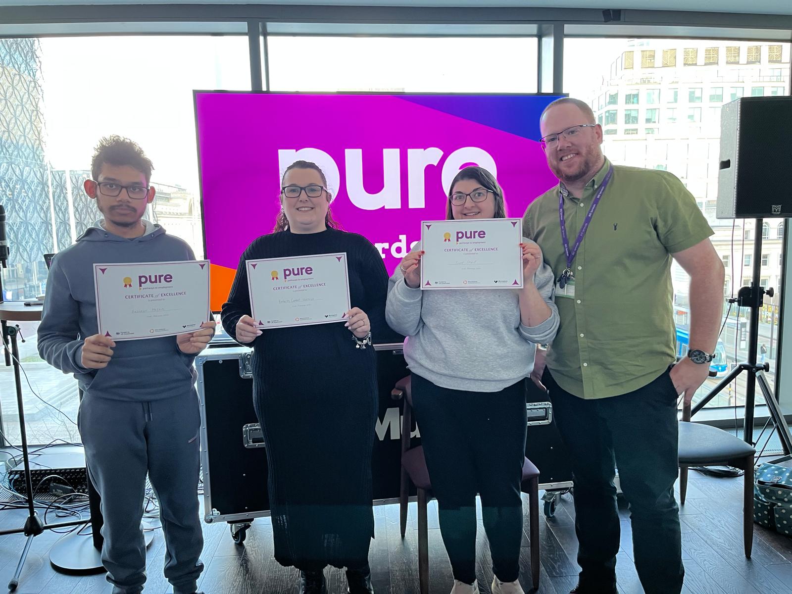 4 people stood side by side smiling towards the camera. A man, a woman and another woman are holding up their individual certificates. To the right of them is a man who is leaning in.