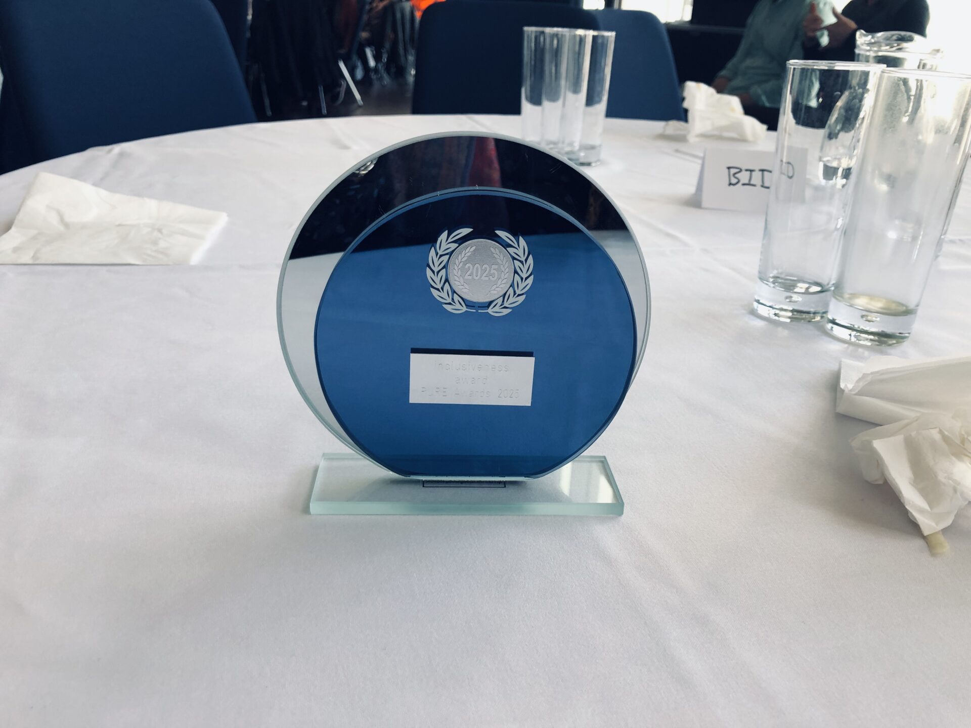 BID's trophy award. Consists of two round mirrored glass pieces stuck together. Near the top is a round silver plaque which reads 2025. The plaque is bordered by symmetrical silver leaves. Below that is a small rectangular silver plaque which reads inclusiveness award. PURE Awards 2025. The trophy is atop a rectangular piece of glass. 